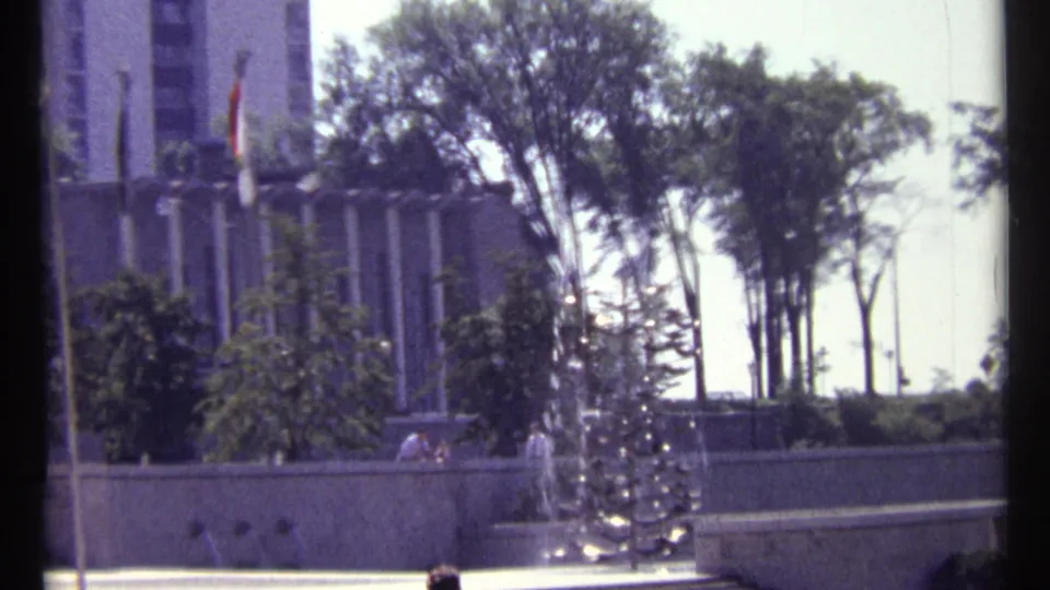1967 Ottawa view of flags, trees, and a spraying public fountain, with distant pedestrians along the edge of the plaza.