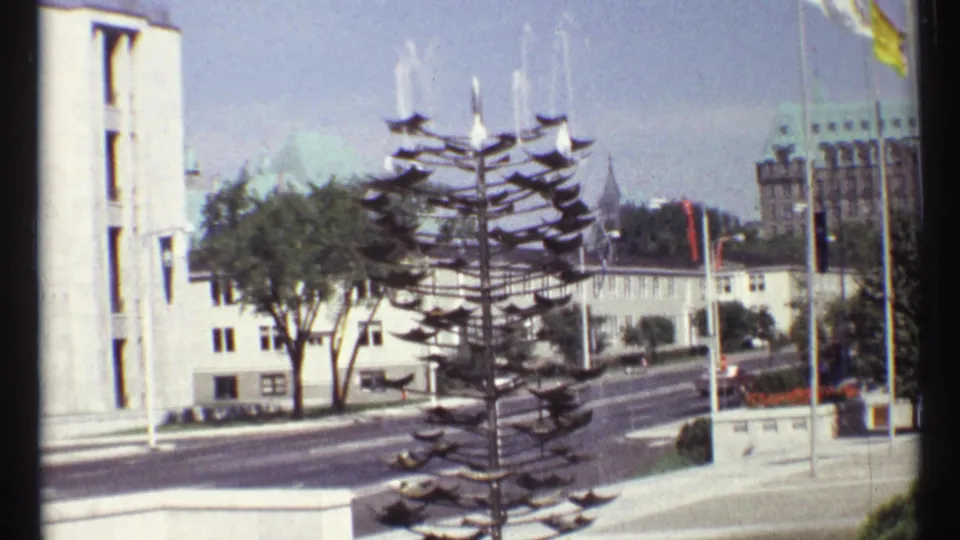 1967 Ottawa home-movie frame showing a sculptural tiered fountain beside a broad street, buildings, flags, trees, and vehicles.
