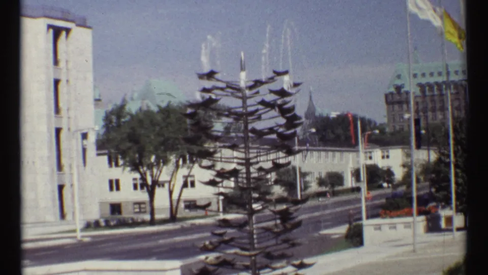1967 Ottawa street view with a tiered fountain in the foreground, flags and buildings beyond, and vehicles on a broad road.
