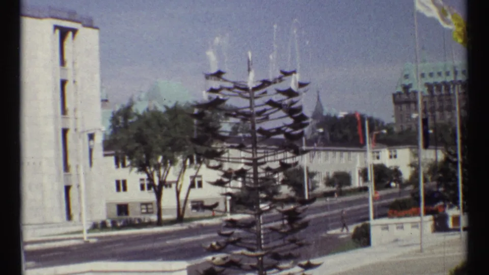 1967 Ottawa street view with a tiered metal sculpture in front of civic buildings, flagpoles, and light traffic.