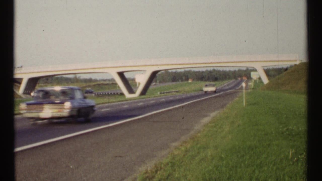 Period cars travel along a highway beneath a concrete overpass beside grassy embankments.