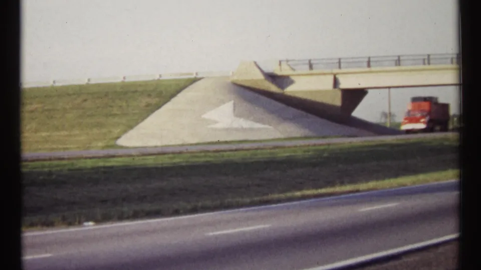 A red truck passes by the concrete overpass on a 1967 Ottawa highway, with embankments and asphalt lanes in view.