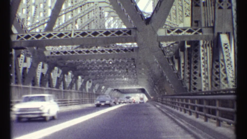 The 1967 Ottawa bridge crossing continues, with a blurred light-colored car passing near the camera under heavy steel trusses.