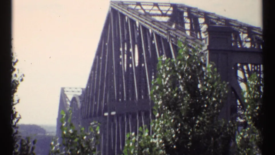 Exterior 1967 Ottawa view of a large steel truss bridge seen past leafy trees and distant water or shoreline.