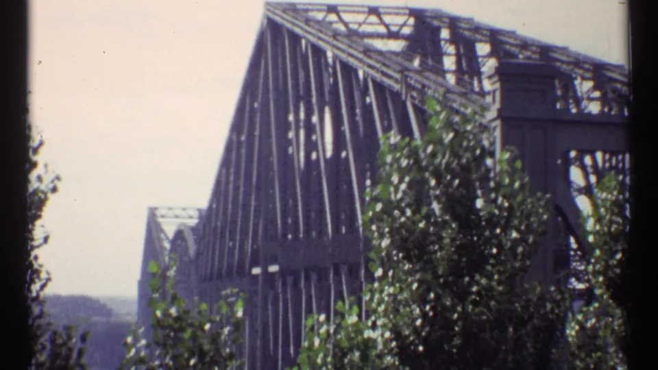 Exterior home-movie frame of the Ottawa steel truss bridge with foreground trees and a hazy background.