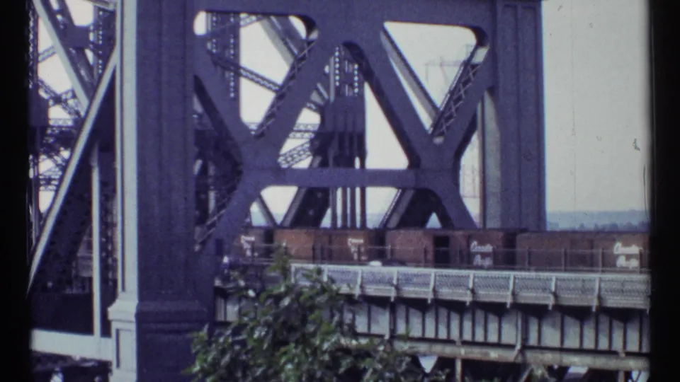 1967 Ottawa 8mm frame showing brown freight cars moving across a large steel truss bridge behind foreground foliage.