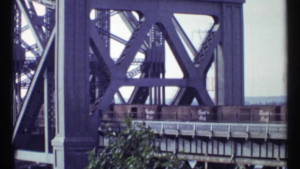 1967 Ottawa frame with freight cars crossing behind heavy geometric steel bridge supports and a leafy foreground edge.