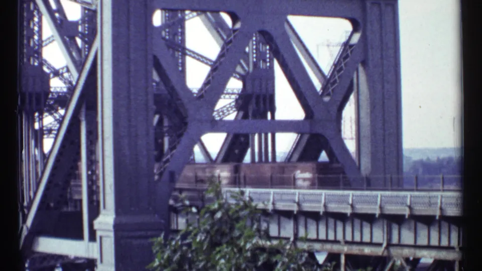 1967 Ottawa 8mm frame of freight cars crossing a large steel bridge, with dark beams and green foliage visible.
