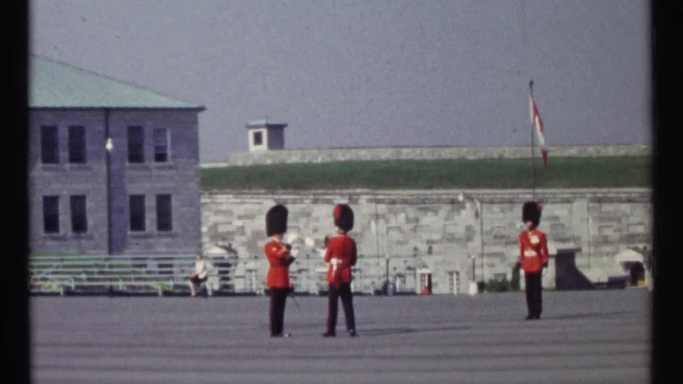 Three ceremonial guards hold drill positions in 1967 Ottawa, framed against a stone wall, green roofline, benches, and a Canadian flag.