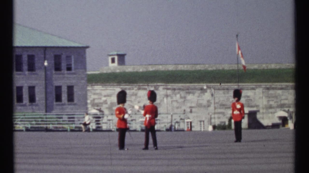 Three guards in red tunics stand on a parade ground near a Canadian flag and stone walls.