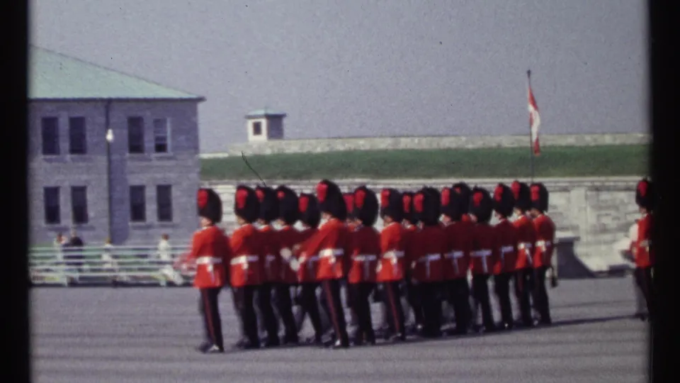 1967 Ottawa footage frame with Canadian guards in red tunics marching past stone buildings and distant onlookers.