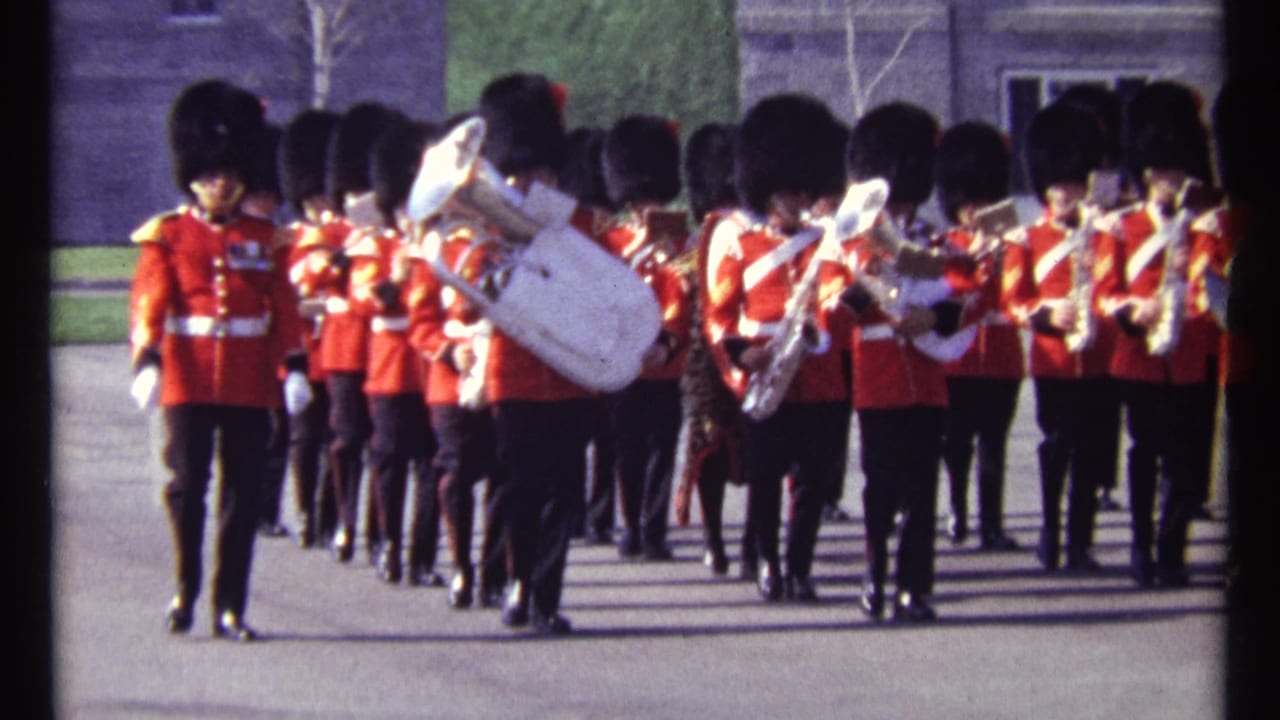 Ceremonial band members in red uniforms advance in formation with a large white tuba near center.