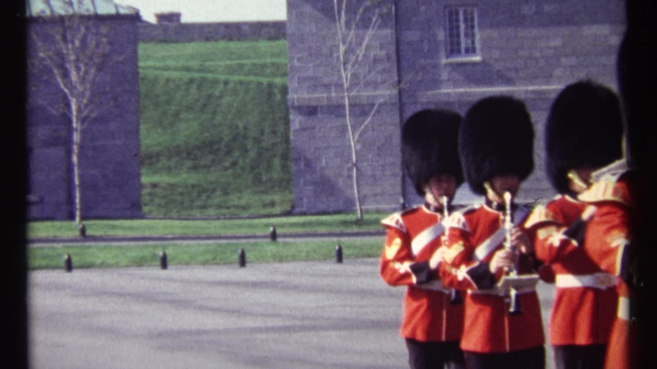 Ottawa 1967 home-movie frame showing uniformed ceremonial musicians in red tunics and black hats before stone buildings and lawn.