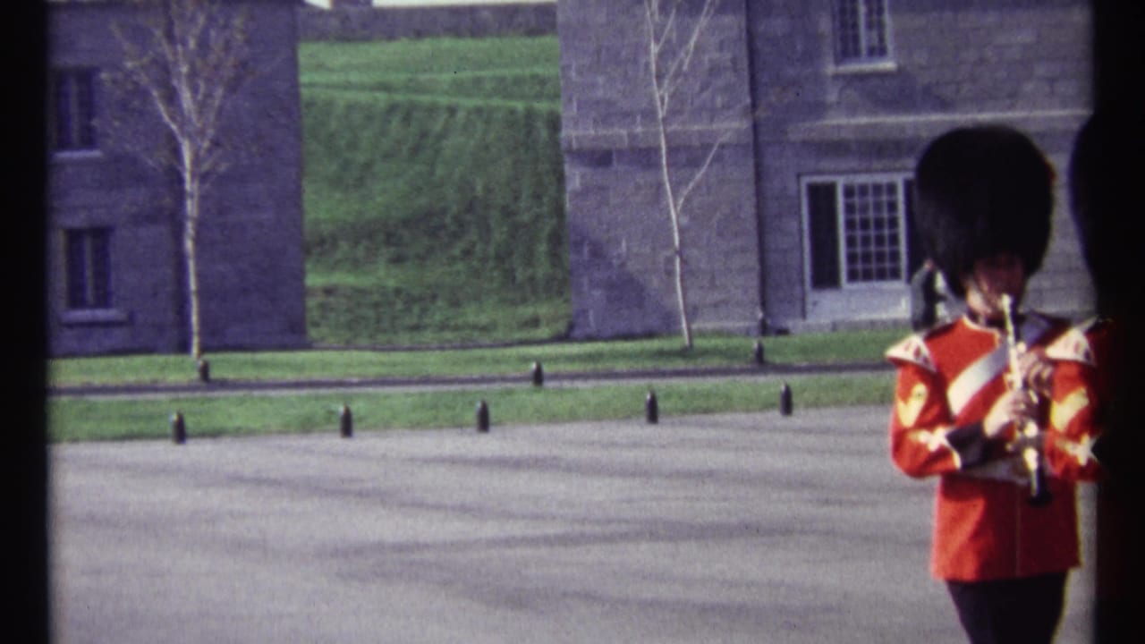 Single uniformed band member in a red tunic and tall black hat holding an instrument on open pavement.