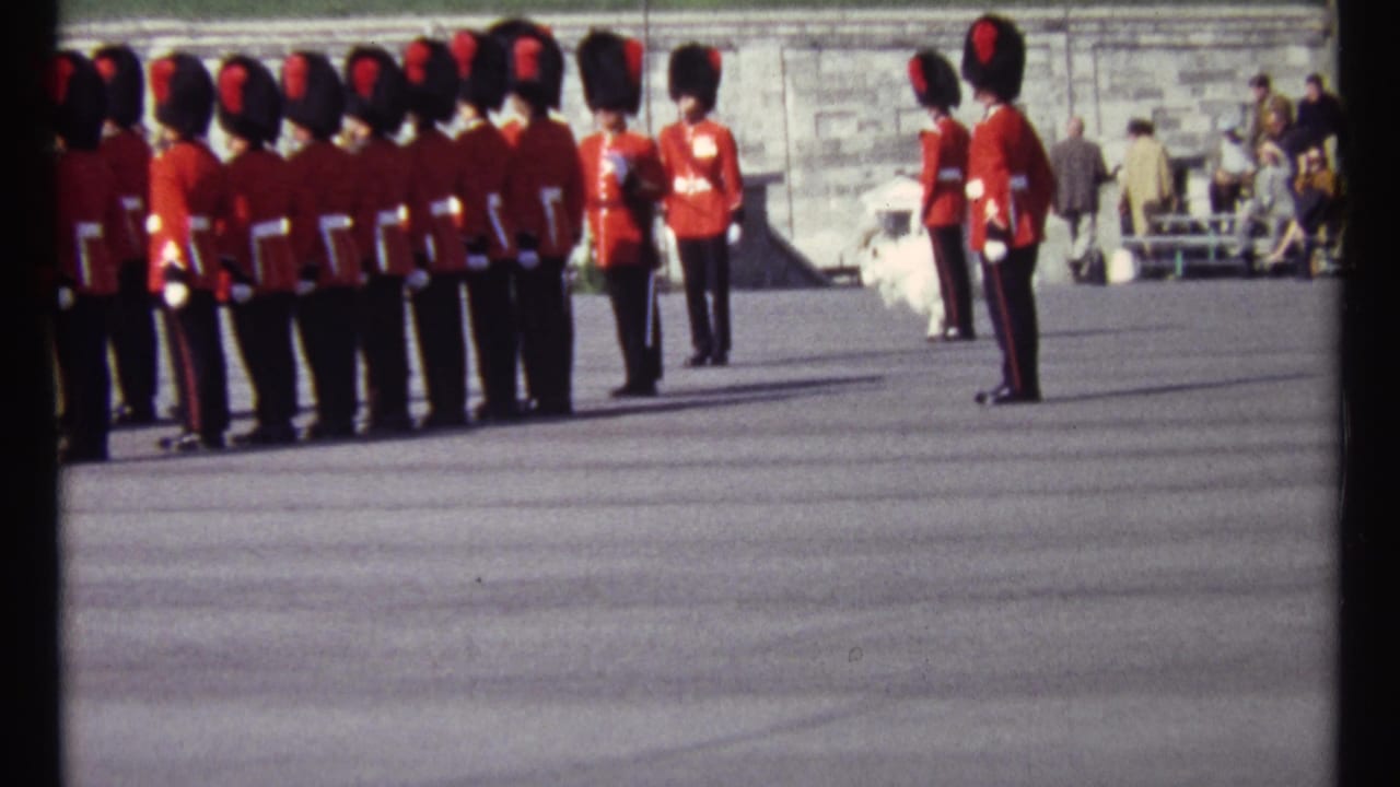 Red-coated guards in tall black hats stand and march across a paved parade ground.