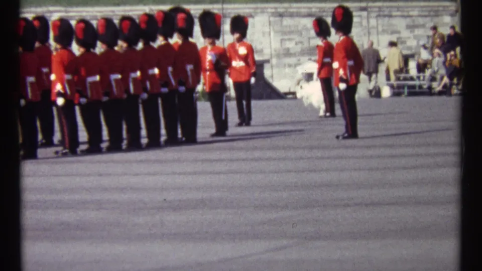 1967 Ottawa home-movie frame showing ceremonial guards in red tunics and tall black hats on a broad paved parade ground.
