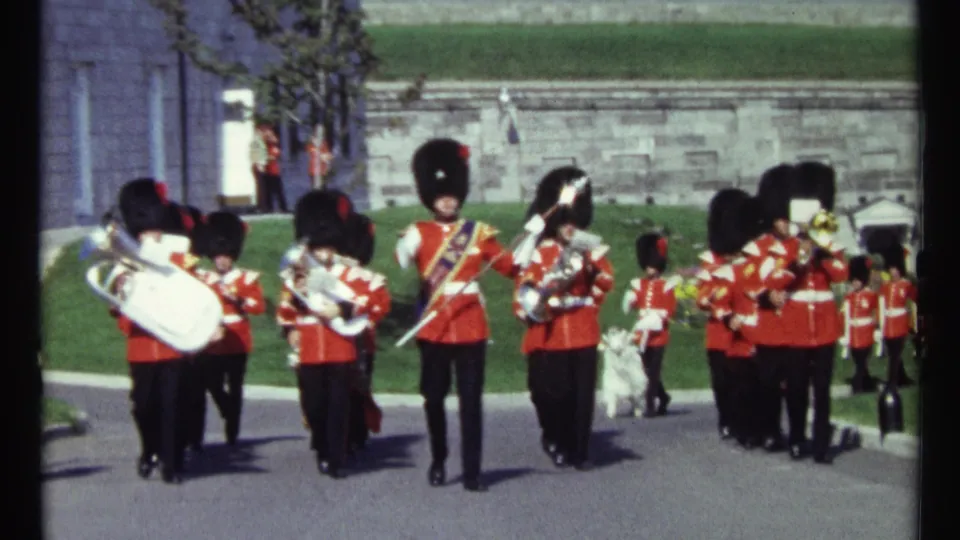 1967 Ottawa home-movie frame showing a ceremonial marching band in red uniforms with brass and drum instruments near stone walls.