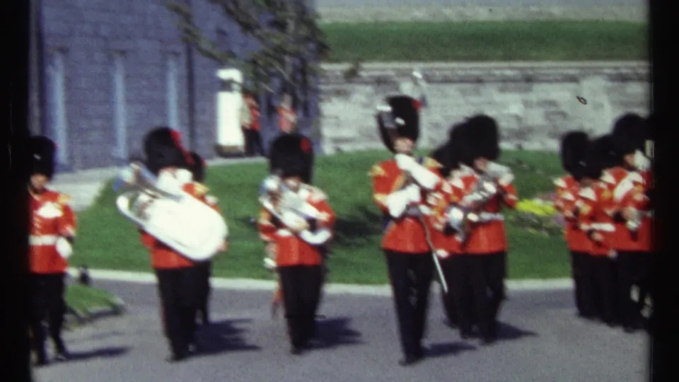 1967 Ottawa ceremonial band scene with red-coated musicians carrying brass instruments and drums beside a lawn and stone wall.