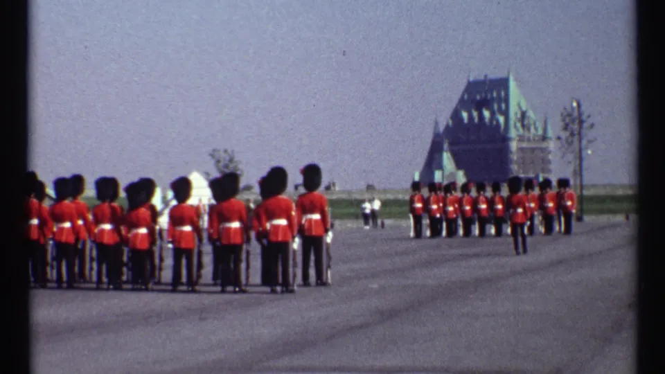 Ottawa ceremonial guards in red tunics and tall black hats stand in ranks across a paved open space with a large roofed building beyond.