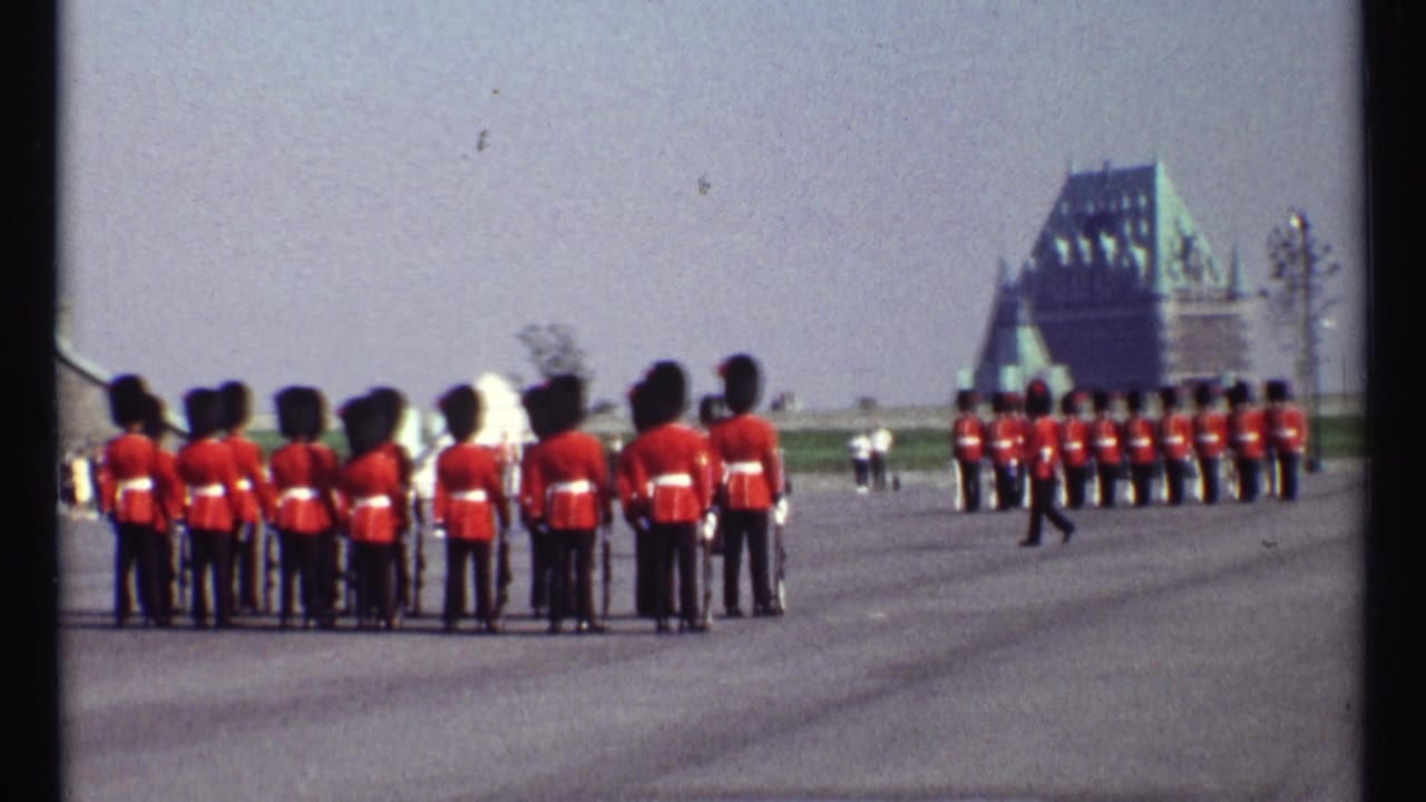 Red-coated guards in bearskin hats stand on a paved parade ground with distant formal buildings.