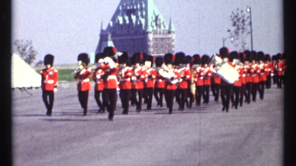 A red-uniformed brass band marches across a broad Ottawa roadway, with a large historic-looking building visible in the background.
