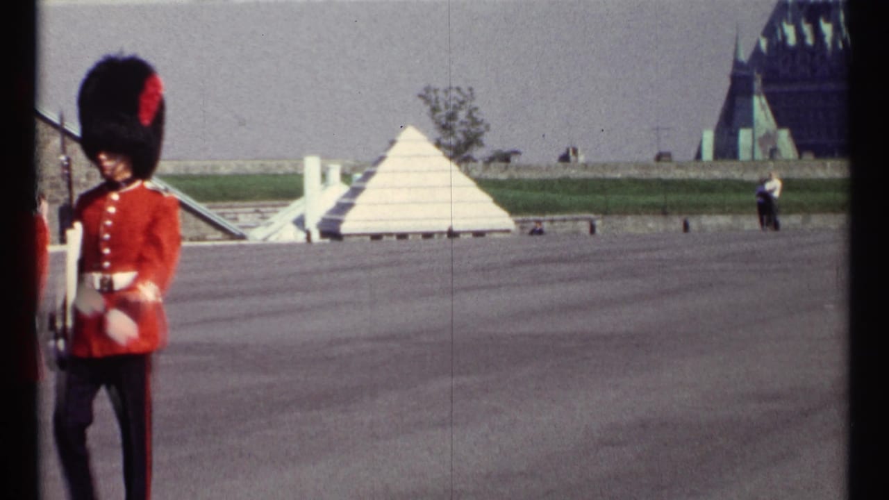 Red-coated guard standing near a wide courtyard with a white pyramid-like structure behind him.