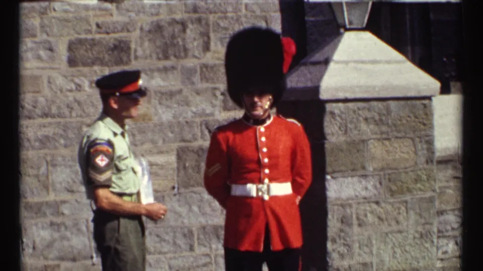 1967 Ottawa 8mm still of an officer and red-coated ceremonial guard beside a textured stone wall and doorway.