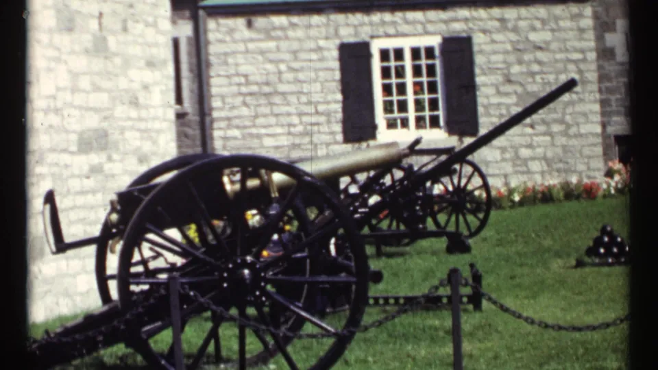 1967 Ottawa home-movie frame showing chained historic cannons on grass beside a stone building with black shutters.