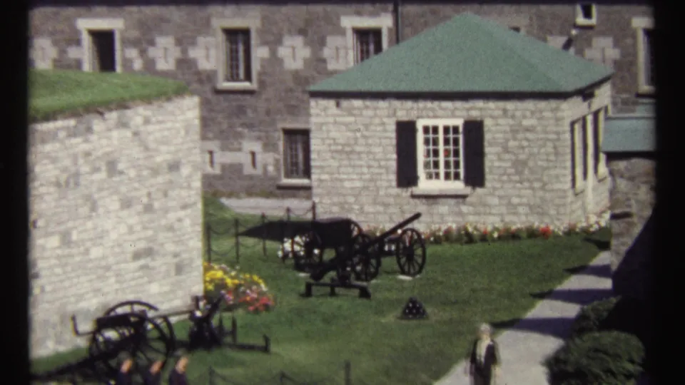 1967 Ottawa home-movie frame showing cannons on a lawn near stone walls and a small green-roofed building with black shutters.