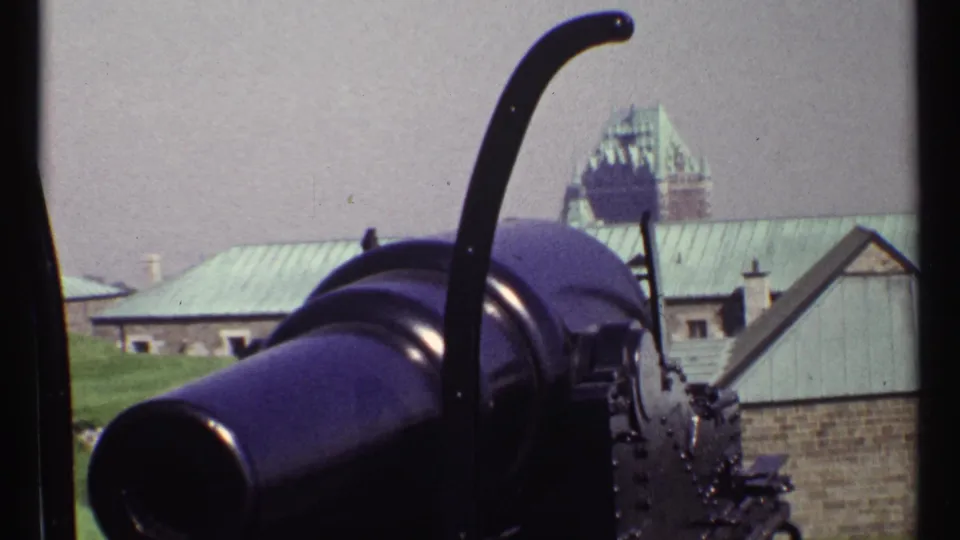 1967 8mm visitor-shot view with a black cannon dominating the foreground and a distant château-style building beyond stone and metal roofs.