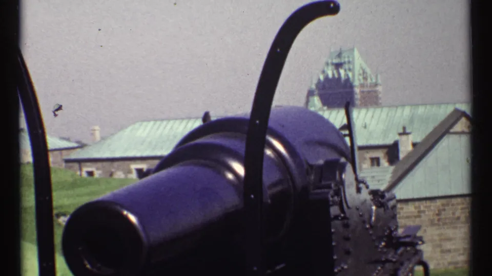 1967 Ottawa home-movie frame showing a close outdoor view of a cannon with stone buildings and green copper roofs in the background.