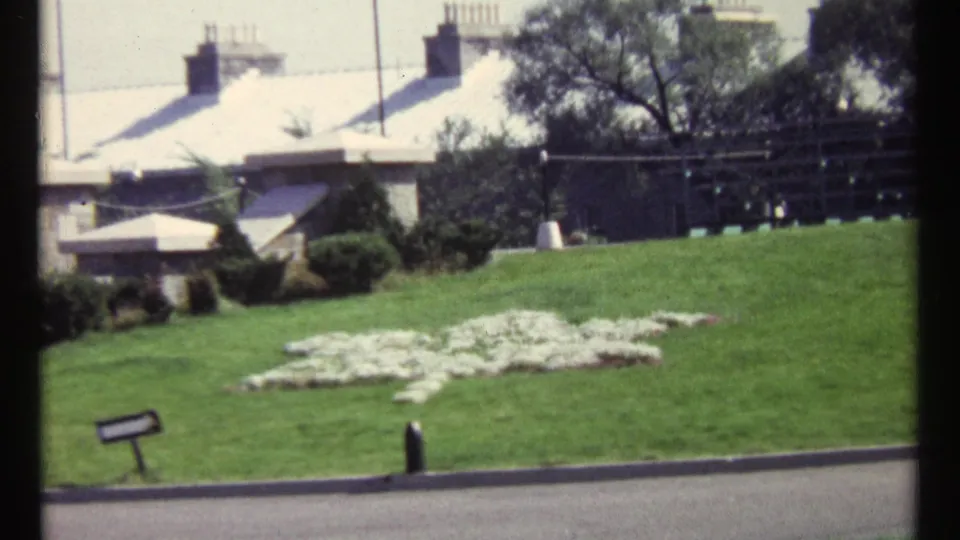 1967 Ottawa home-movie frame with a white peace-symbol flower arrangement on a sloped lawn near a small sign and background buildings.