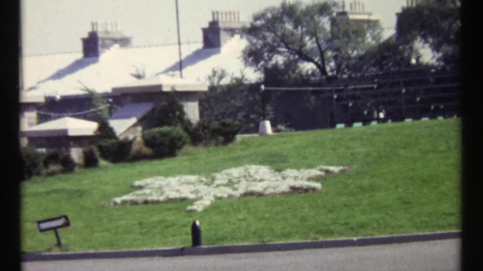 1967 Ottawa frame showing the peace-symbol flower bed from a similar angle, with lawn, sign, trees, and rooftops visible behind it.