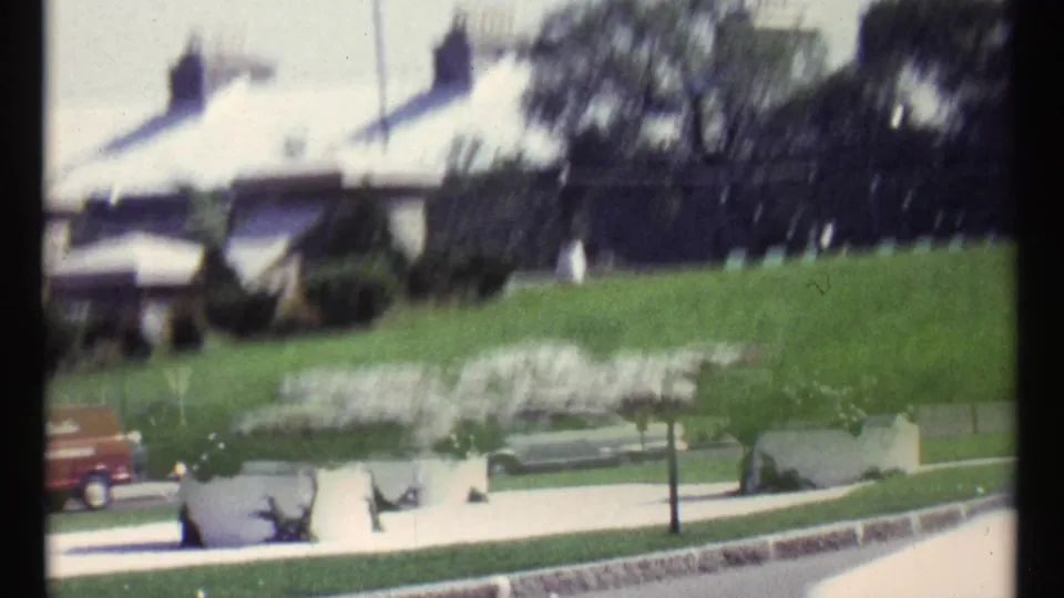 1967 Ottawa home-movie frame showing cars near a tree-lined street, landscaped planters, grass, and a pale building in bright daylight.