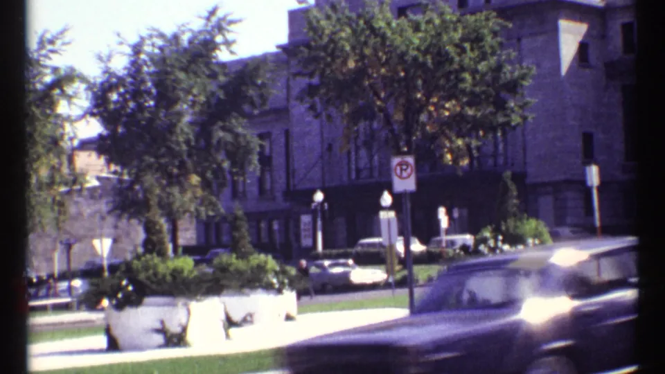 1967 Ottawa frame with a dark car crossing the foreground, pedestrians near planters, trees, stone buildings, and street signage.