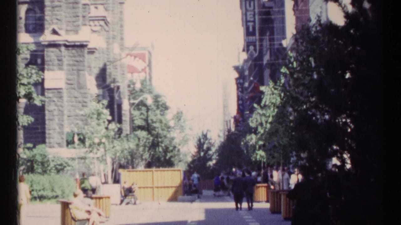 Sunny sidewalk scene with pedestrians, benches, trees, stone building, and a vertical commercial sign.