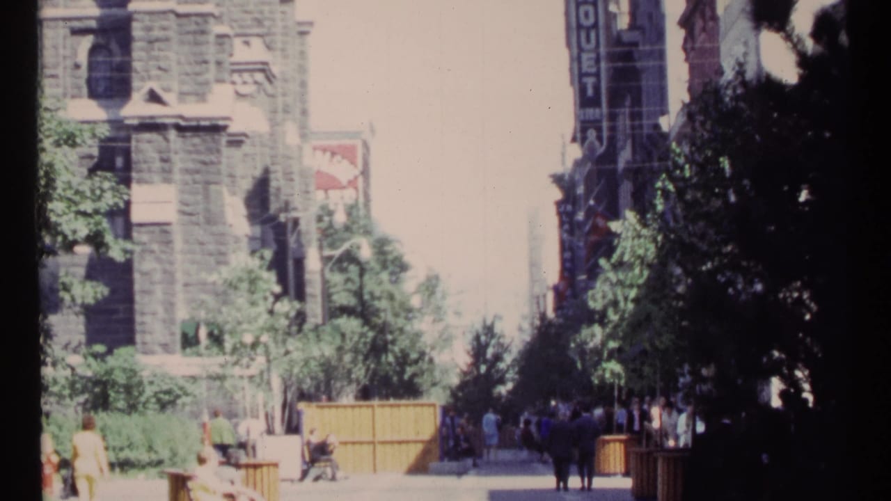 Pedestrians walk and sit along a tree-lined sidewalk beside stone buildings and wooden barriers.