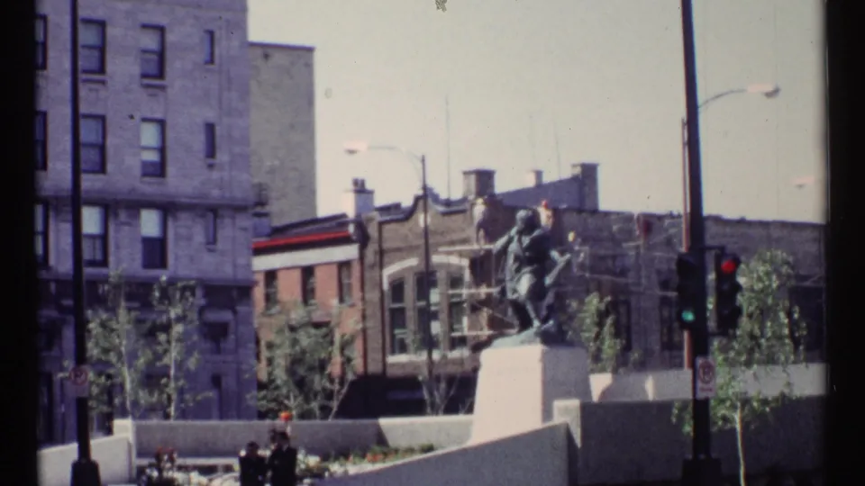 Ottawa home-movie frame showing a statue on a pedestal with mid-century downtown buildings and street fixtures behind it.