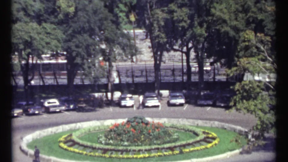 Ottawa home-movie frame from 1967 showing a circular landscaped bed with parked cars and trees beyond.