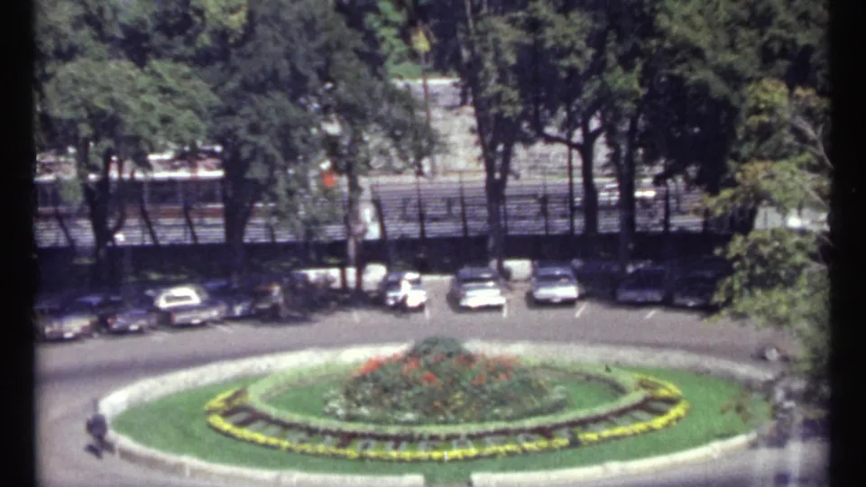 Elevated 1967 Ottawa view of a formal roundabout garden, parked mid-century cars, trees, and distant pedestrians.
