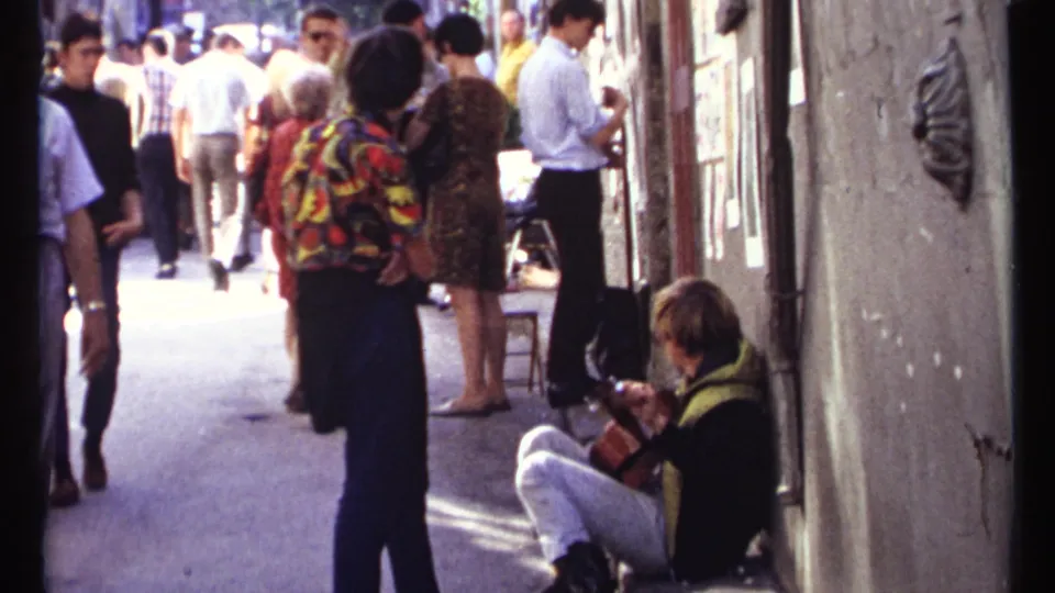 1967 Ottawa footage frame showing a busy walkway, people in period clothing, exterior display panels, and a guitarist seated against the wall.
