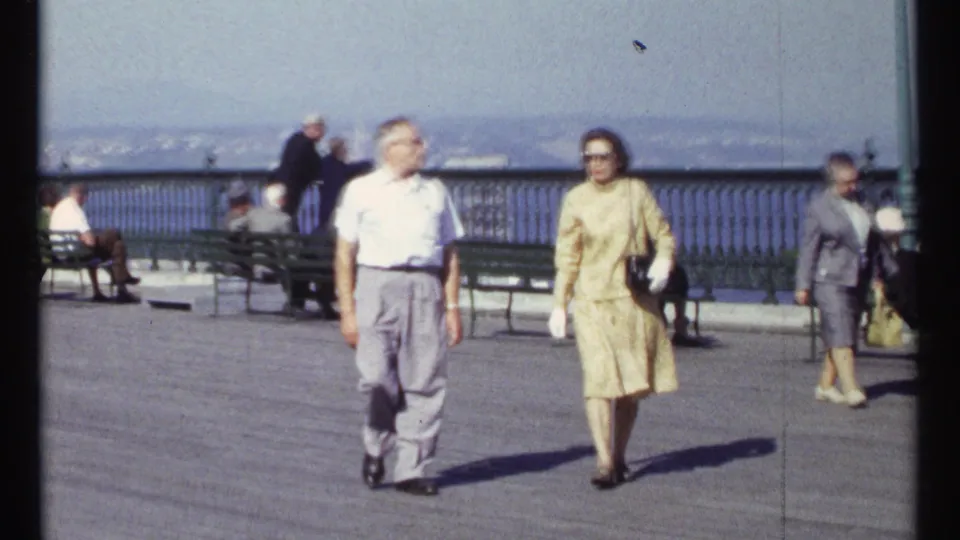 Ottawa home-movie frame from 1967 showing pedestrians walking along a waterfront promenade with benches, railing, and water beyond.