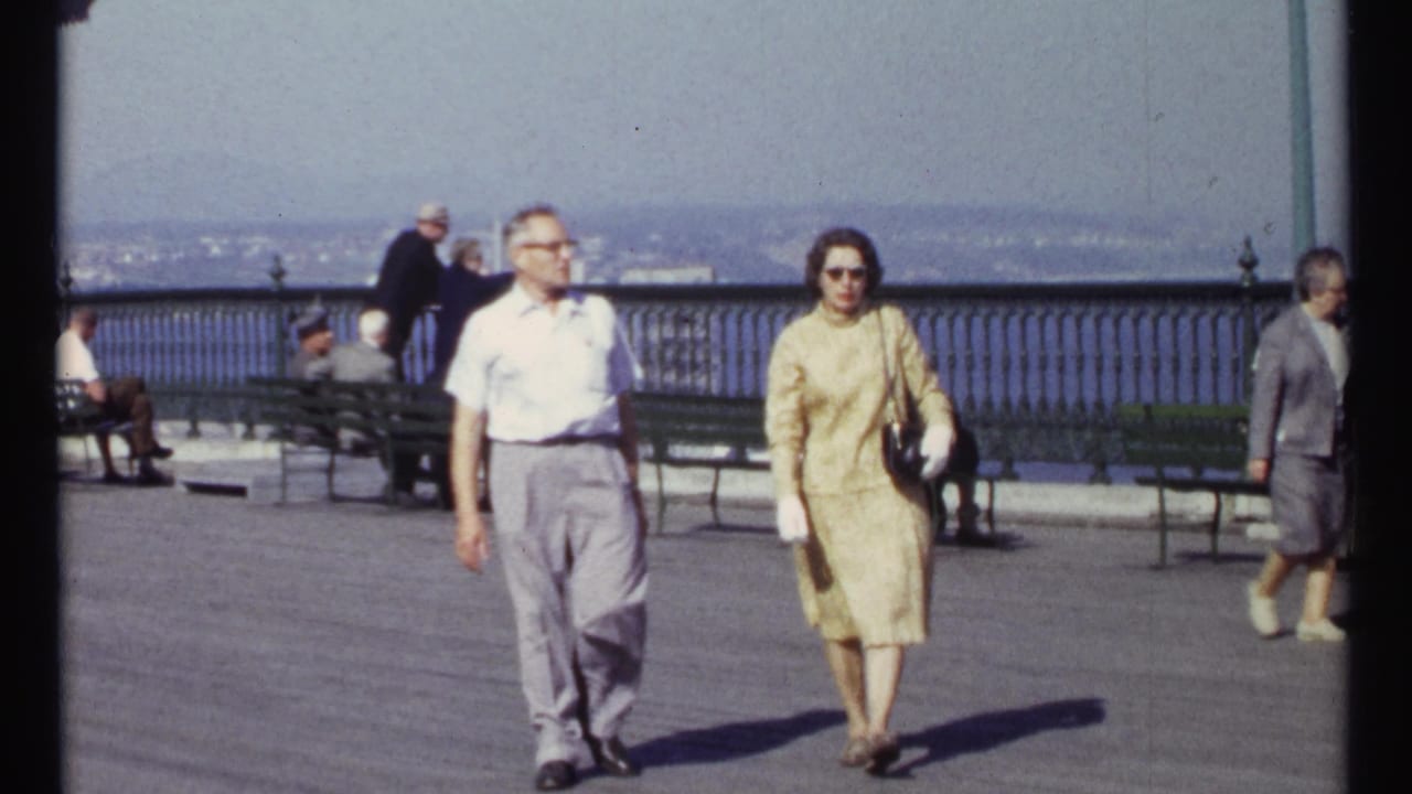 A man and woman walk on a paved promenade with benches, railing, water, and distant shoreline behind them.