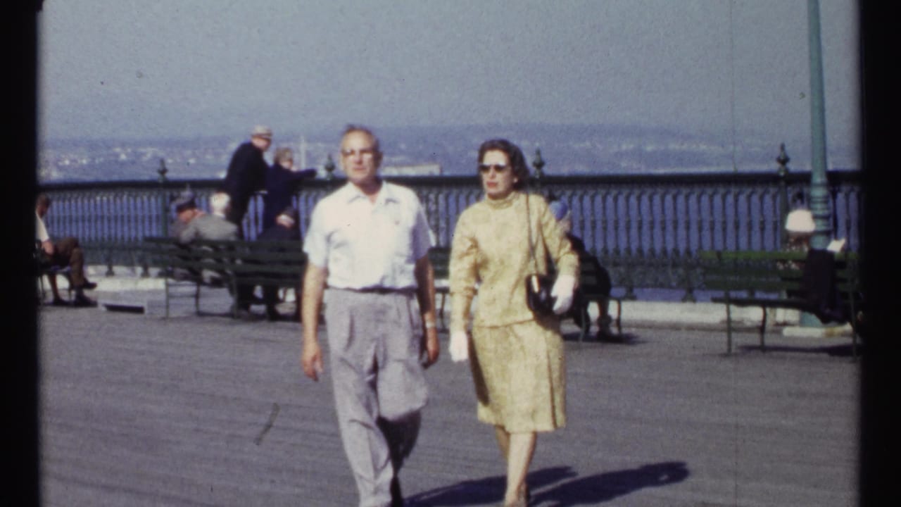 The couple continues forward on the promenade, with seated visitors, railing, water, and a distant skyline behind them.