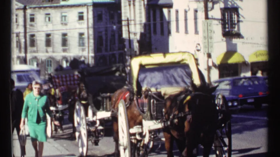 A woman in a green dress walks by horse-drawn buggies on a sunny 1967 Ottawa street with cars behind her.