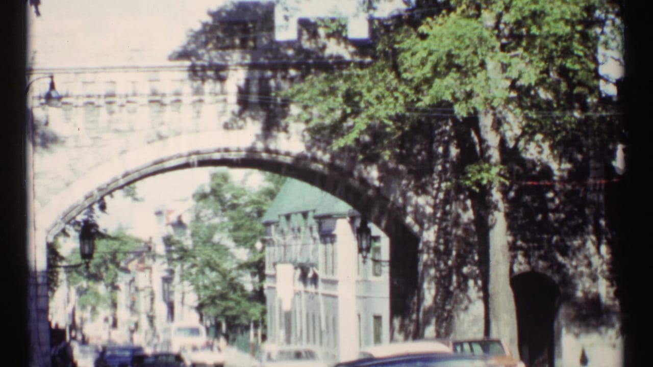 Stone arch bridge over a city street with cars, trees, lamps, and pedestrians.