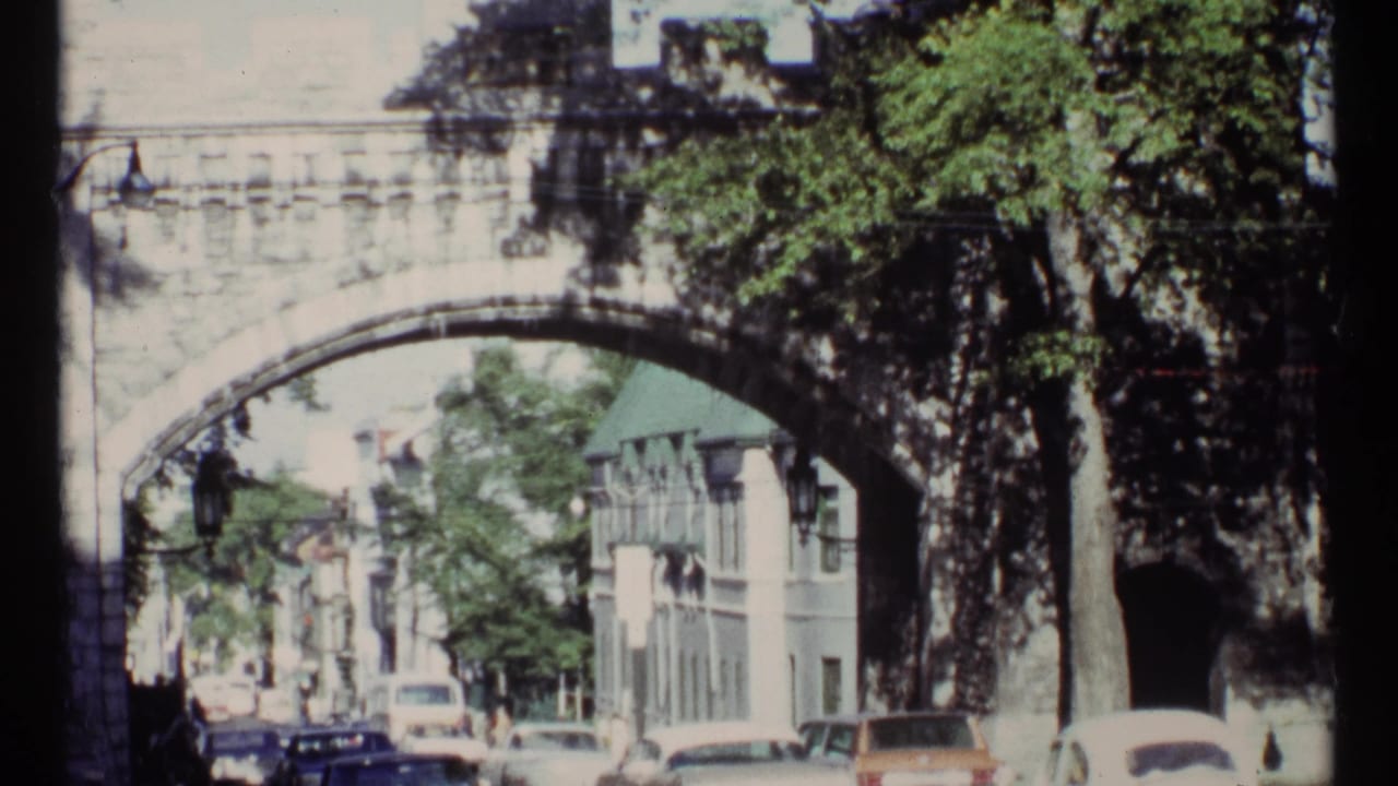 1967 Ottawa view of cars and pedestrians passing under a stone arch bridge beside trees and older buildings.