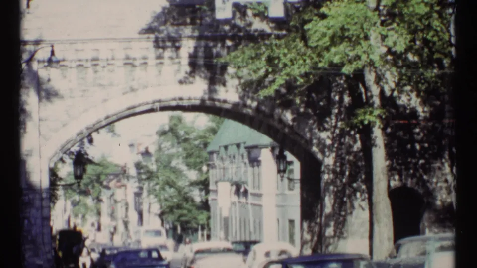 1967 Ottawa home-movie frame of a stone arch bridge over a street with classic cars, pedestrians, trees, and nearby buildings.