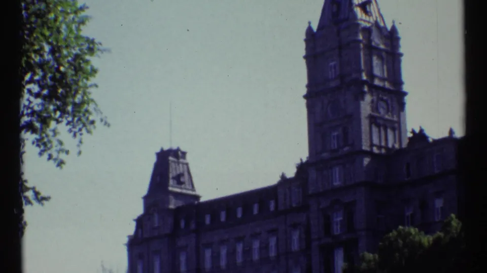 1967 Ottawa home-movie frame showing Château Laurier's stone tower, steep roofline, leafy branches, and pale sky.