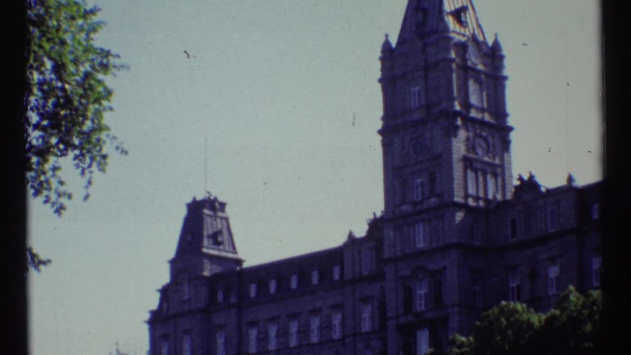 Stone hotel facade with tall tower, mansard roofline, trees, and pale sky.